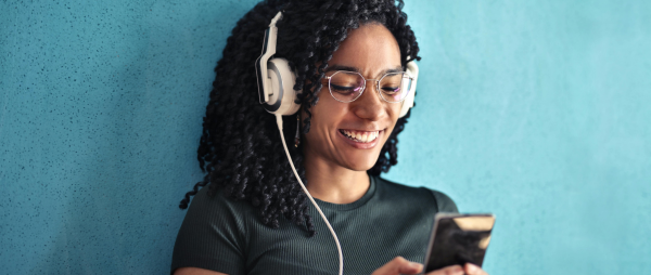 A smiling woman looks at her phone, she has black hair, glasses and is wearing large white and grey headphones.