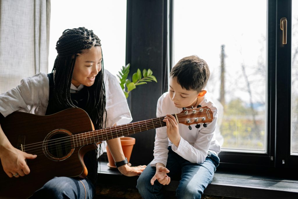 A young girl is holding a guitar, a young boy sits with his hand over the fretboard. It looks as though she is teaching him the instrument.