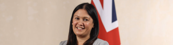 An image of Lisa Nandy smiling at the camera. She has black hair and is wearing a grey top. The flag of the United Kingdom is behind her.