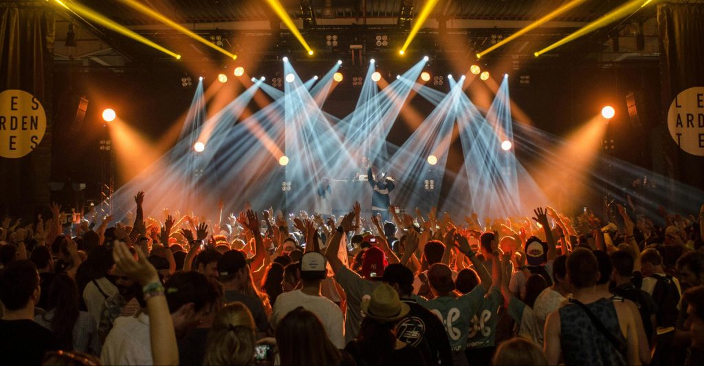 A photo of a brightly lit stage, there is a clapping audience in the foreground and artists performing in the background.